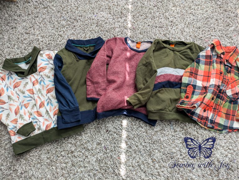 White with leaves sweater, dark green and blue sweater, red and blue sweater, green sweater with gray red and blue rainbow across front, and orange and green button up are on carpet background.